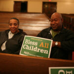 photo of couple sitting in church holding sign that says, "bless all children"
